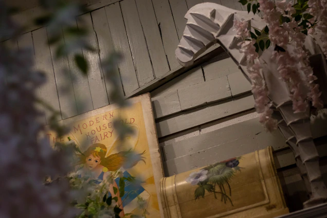 A vintage-style book titled 'The Modern Household Fairy' is partially obscured by floral arrangements, set against a backdrop of wooden paneling. A sculpture resembling a horse is adorned with pink flowers.