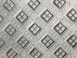 An intricate pattern of embossed diamond shapes on a textured surface, with each diamond containing a cross-like design inside. The texture resembles carved stone or concrete, giving it a three-dimensional appearance.