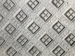 An intricate pattern of embossed diamond shapes on a textured surface, with each diamond containing a cross-like design inside. The texture resembles carved stone or concrete, giving it a three-dimensional appearance.
