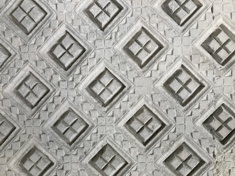 An intricate pattern of embossed diamond shapes on a textured surface, with each diamond containing a cross-like design inside. The texture resembles carved stone or concrete, giving it a three-dimensional appearance.