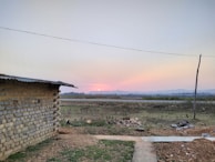 A rural landscape at sunset with a small brick building on the left, an open field in the center and background, and a distant mountain range beneath a colorful sky. A rooster is visible near the building, and a single electrical wire runs across the scene.