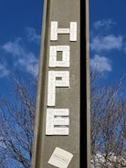A vertical concrete structure with electronic doorbell buttons arranged to spell out a word, set against a clear blue sky with some clouds. Leafless tree branches are visible in the background. There's a small paper or sticker at the bottom with text.