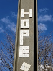 A vertical concrete structure with electronic doorbell buttons arranged to spell out a word, set against a clear blue sky with some clouds. Leafless tree branches are visible in the background. There's a small paper or sticker at the bottom with text.