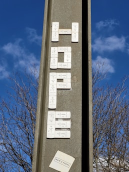 A vertical concrete structure with electronic doorbell buttons arranged to spell out a word, set against a clear blue sky with some clouds. Leafless tree branches are visible in the background. There's a small paper or sticker at the bottom with text.