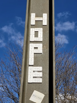A vertical concrete structure with electronic doorbell buttons arranged to spell out a word, set against a clear blue sky with some clouds. Leafless tree branches are visible in the background. There's a small paper or sticker at the bottom with text.