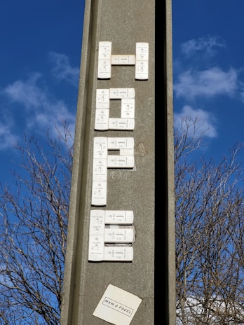A vertical concrete structure with electronic doorbell buttons arranged to spell out a word, set against a clear blue sky with some clouds. Leafless tree branches are visible in the background. There's a small paper or sticker at the bottom with text.