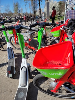 Several Lime-branded scooters and bicycles are parked on a paved area. These are accompanied by a group of people in the background, some walking and others gathered around. The image shows a clear, sunny day with trees having white blossoms and light green leaves.