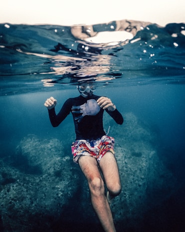 A person wearing colorful shorts and a black long-sleeve shirt is snorkeling underwater. They are holding a jellyfish, and their face is partly submerged but visible through a snorkeling mask. The water is clear, with a rocky seabed visible.