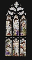 An intricately designed stained glass window depicting biblical scenes with figures in traditional garments. Angels are portrayed at the top section, and the lower panels show various scenes involving biblical characters with halos and vibrant robes. The artwork features detailed expressions and symbolic elements like staffs and scrolls.