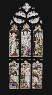An intricately designed stained glass window depicting biblical scenes with figures in traditional garments. Angels are portrayed at the top section, and the lower panels show various scenes involving biblical characters with halos and vibrant robes. The artwork features detailed expressions and symbolic elements like staffs and scrolls.