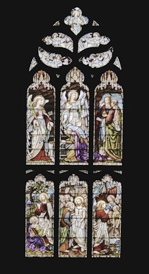 An intricately designed stained glass window depicting biblical scenes with figures in traditional garments. Angels are portrayed at the top section, and the lower panels show various scenes involving biblical characters with halos and vibrant robes. The artwork features detailed expressions and symbolic elements like staffs and scrolls.