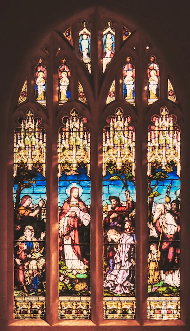 Detailed stained glass window depicting religious figures, with intricate designs and vibrant colors in a church setting. The window features biblical scenes and characters surrounded by ornate patterns.