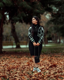 A woman standing in a park surrounded by fallen autumn leaves, wearing a black dress, checkered jacket, and sneakers. She holds a small black purse, and the background is filled with green trees and blurred scenery.