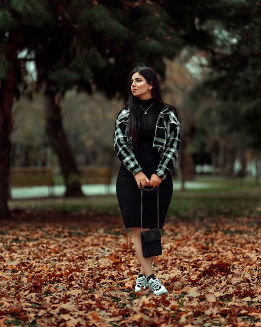 A woman standing in a park surrounded by fallen autumn leaves, wearing a black dress, checkered jacket, and sneakers. She holds a small black purse, and the background is filled with green trees and blurred scenery.