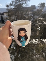 A hand holding a white mug with a small polka dot pattern featuring a figurine of a nurse wearing teal scrubs and a stethoscope. The background is an outdoor setting with leafy trees and a view of buildings under a clear sky.
