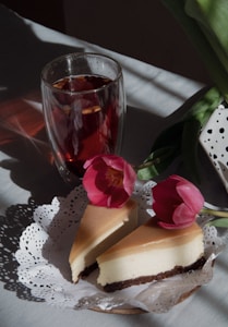 A glass of dark red tea with floating ingredients sits beside a plate with two slices of cheesecake decorated with vibrant pink tulips. The setup rests on a white doily on a light fabric, with sunlight casting shadows across the scene.