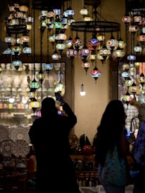 A vibrant display of multicolored mosaic lamps hanging from the ceiling in a dimly lit shop. Silhouetted figures stand in the foreground, with one person capturing a photo with a phone. The shop interior is filled with various intricately designed ceramics and artifacts, creating an exotic and artistic ambiance.