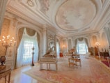 A luxurious, ornately decorated room features intricate architectural details on the ceiling and walls, with large draped windows allowing natural light to filter in. A grand piano is positioned centrally next to elegant antique furniture arranged on a lush, patterned rug. The room is illuminated by multiple classic chandeliers, adding to the opulent ambiance.