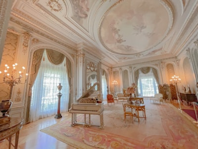 A luxurious, ornately decorated room features intricate architectural details on the ceiling and walls, with large draped windows allowing natural light to filter in. A grand piano is positioned centrally next to elegant antique furniture arranged on a lush, patterned rug. The room is illuminated by multiple classic chandeliers, adding to the opulent ambiance.