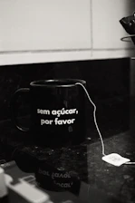 A black mug with the phrase 'sem açúcar, por favor' written on it sits on a dark countertop. A tea bag with a string dangles over the edge, and the tag is visible next to the mug.