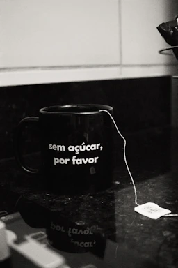 A black mug with the phrase 'sem açúcar, por favor' written on it sits on a dark countertop. A tea bag with a string dangles over the edge, and the tag is visible next to the mug.