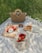 A picnic setup on a light-colored blanket, featuring a wicker bag with round handles, a container of strawberries in a netted bag, a round of cheese with cherry tomatoes, a jar of spread, and a pouch containing baguette pieces. The scene takes place on a grass-covered area with dappled sunlight filtering through.
