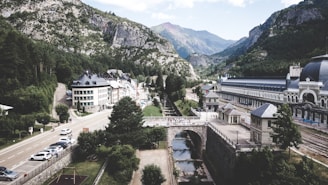 A picturesque mountain village features a mixture of modern and traditional buildings lining a narrow street, flanked by lush green trees and set against a backdrop of majestic mountains. A river runs alongside a railway station, complete with arched bridges and tracks, indicating a historical or possibly a touristic area.