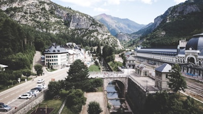 A picturesque mountain village features a mixture of modern and traditional buildings lining a narrow street, flanked by lush green trees and set against a backdrop of majestic mountains. A river runs alongside a railway station, complete with arched bridges and tracks, indicating a historical or possibly a touristic area.