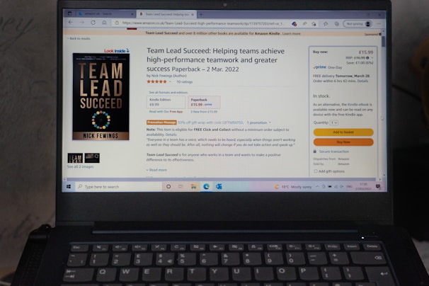 A laptop displays an Amazon product page for the book 'Team Lead Succeed'. The book cover is visible on the left, highlighting the title in bold letters. The page shows options to purchase in paperback or Kindle format, with price details and shipping options. The background includes a promotional message and a quote from the book.
