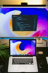 A workspace featuring a large desktop monitor displaying a vibrant abstract wallpaper with a code editor open, and a laptop with the same wallpaper in front. The desk setup includes a keyboard, mouse, headphones, a small potted plant, and an AirPods case. The scene conveys a modern, tech-focused environment.