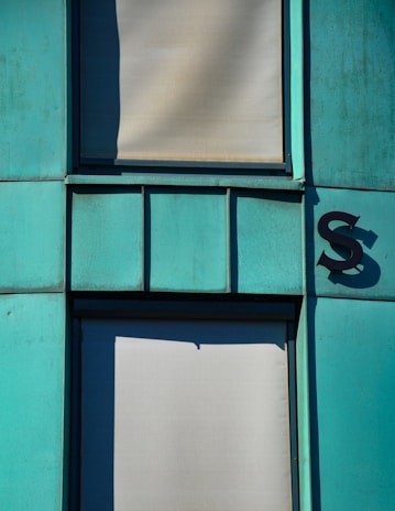 The image features a section of a building façade with turquoise-colored panels and two shaded windows covered by blinds. There is a shadow creating an interesting geometric shape on one of the panels. A large letter 'S' is affixed to the right side of the paneling, casting its own shadow.