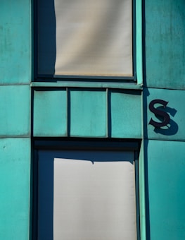 The image features a section of a building fa&ccedil;ade with turquoise-colored panels and two shaded windows covered by blinds. There is a shadow creating an interesting geometric shape on one of the panels. A large letter 'S' is affixed to the right side of the paneling, casting its own shadow.