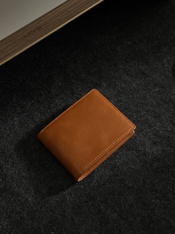 A brown leather wallet is placed on a dark textured surface. The lighting accentuates the smooth texture and stitching of the wallet, adding a touch of elegance. In the corner, part of a wooden object with an inscription is visible.