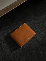 A brown leather wallet is placed on a dark textured surface. The lighting accentuates the smooth texture and stitching of the wallet, adding a touch of elegance. In the corner, part of a wooden object with an inscription is visible.