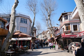 A lively street scene features various small businesses such as restaurants and cafes with awnings. People are walking and sitting at outdoor tables, enjoying the sunny day. Leafless trees line the street, and the sky is clear and blue.