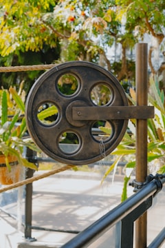 A large metal pulley with several circular cutouts is mounted on a vertical pole. It is part of a mechanism that includes a thick rope threaded through it. The background features lush green foliage and plants, suggesting a garden or outdoor setting.