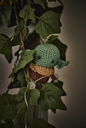 A crocheted green character with large black eyes and small ears is nestled among a set of lush green leaves. The character appears to be in a playful pose, blending in with the surrounding foliage. The lighting is dim, giving a slightly mystical ambiance to the scene.