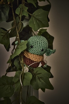 A crocheted green character with large black eyes and small ears is nestled among a set of lush green leaves. The character appears to be in a playful pose, blending in with the surrounding foliage. The lighting is dim, giving a slightly mystical ambiance to the scene.