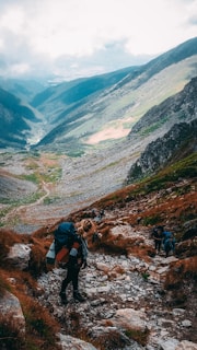 A group of hikers ascends a rocky mountain trail surrounded by breathtaking views of a verdant valley and distant mountains under a cloudy sky. The terrain is rough, with patches of green grass and brown rocks.