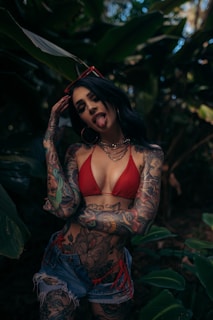 A person with extensive tattoos poses playfully in a jungle-like setting, wearing a red bikini top and denim shorts. Large green leaves surround the individual, who is sticking out their tongue and wearing red-framed sunglasses on their head.
