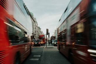 A busy urban street with red double-decker buses in motion, creating a sense of movement and hustle. Historical buildings line the street under an overcast sky, and multiple vehicles, including black cabs, are present on the road.