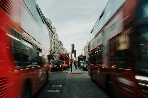 A busy urban street with red double-decker buses in motion, creating a sense of movement and hustle. Historical buildings line the street under an overcast sky, and multiple vehicles, including black cabs, are present on the road.