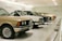 A row of classic cars displayed in a spacious showroom with a sleek, modern design. The cars vary in color and model, featuring vintage designs and pristine conditions, indicating a curated collection.