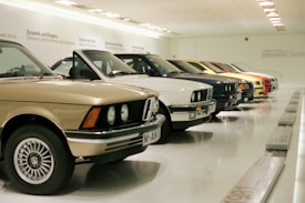 A row of classic cars displayed in a spacious showroom with a sleek, modern design. The cars vary in color and model, featuring vintage designs and pristine conditions, indicating a curated collection.
