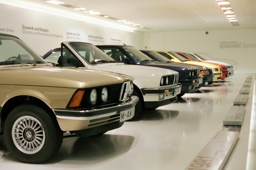 A row of classic cars displayed in a spacious showroom with a sleek, modern design. The cars vary in color and model, featuring vintage designs and pristine conditions, indicating a curated collection.