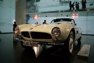 A classic white convertible car is on display in an indoor showroom. The car features round headlights, a distinctive front grille, and a license plate with the code MAE 507H. The showroom has high ceilings with visible lighting fixtures and walls made of glass, allowing silhouettes of people to be seen in the background.