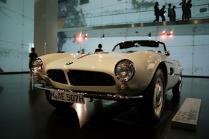 A classic white convertible car is on display in an indoor showroom. The car features round headlights, a distinctive front grille, and a license plate with the code MAE 507H. The showroom has high ceilings with visible lighting fixtures and walls made of glass, allowing silhouettes of people to be seen in the background.