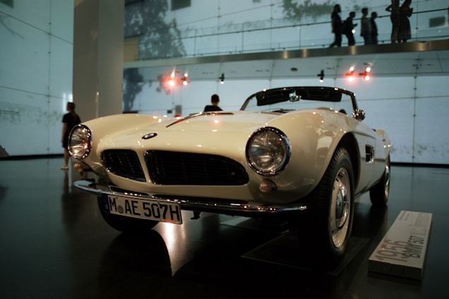 A classic white convertible car is on display in an indoor showroom. The car features round headlights, a distinctive front grille, and a license plate with the code MAE 507H. The showroom has high ceilings with visible lighting fixtures and walls made of glass, allowing silhouettes of people to be seen in the background.