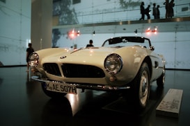 A classic white convertible car is on display in an indoor showroom. The car features round headlights, a distinctive front grille, and a license plate with the code MAE 507H. The showroom has high ceilings with visible lighting fixtures and walls made of glass, allowing silhouettes of people to be seen in the background.