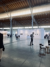 An art exhibition in a large, spacious hall with high ceilings, illuminated by soft lighting from the windows above. White walls partition the space into different sections, each displaying various artworks. Several people are strolling around, observing the art. A few chairs are scattered across the floor.